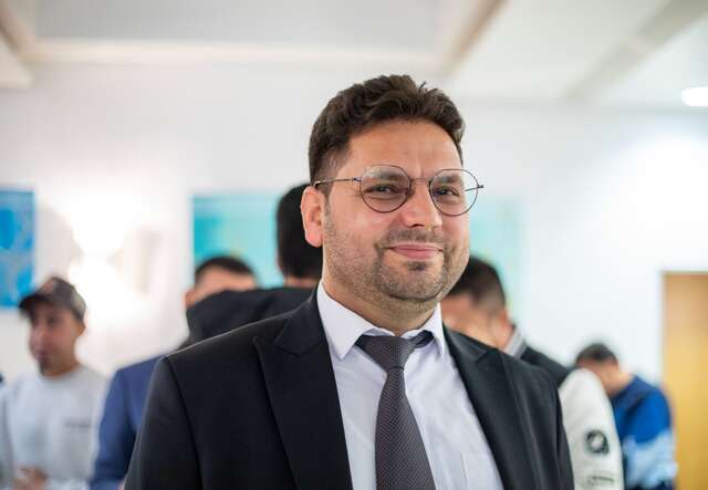 Ein Mann in einem Anzug und mit Brille steht in einem Raum, umgeben von anderen Menschen.